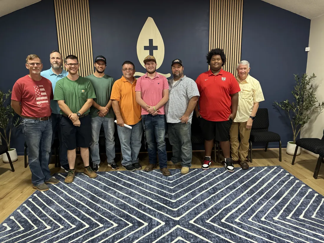 Nine men of every age standing together beneath the flame-and-cross wall at The Landing after the first Forge breakfast